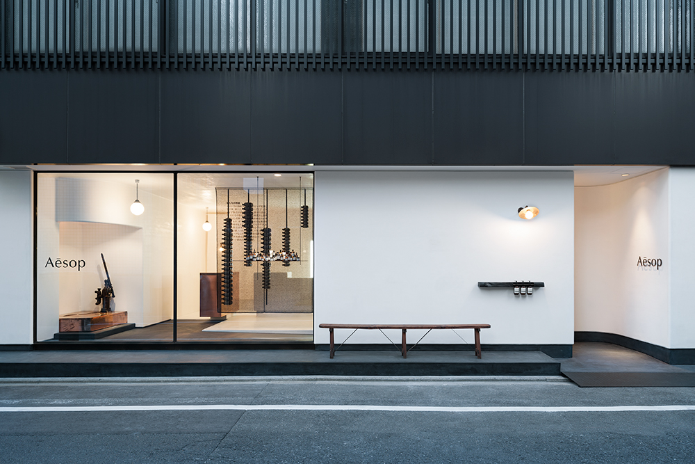 En noir et blanc, rehaussée d’accents de cuivre, la boutique de Kyoto créée par le bureau de Shinichiro Ogata, Simplicity, est empreinte de la culture japonaise du 14ème siècle à aujourd’hui. Des voiles en mailles noircies dans lesquelles sont cousues des bouteilles Aesop organisent l’espace dans sa verticalité comme autant de pages calligraphiées.