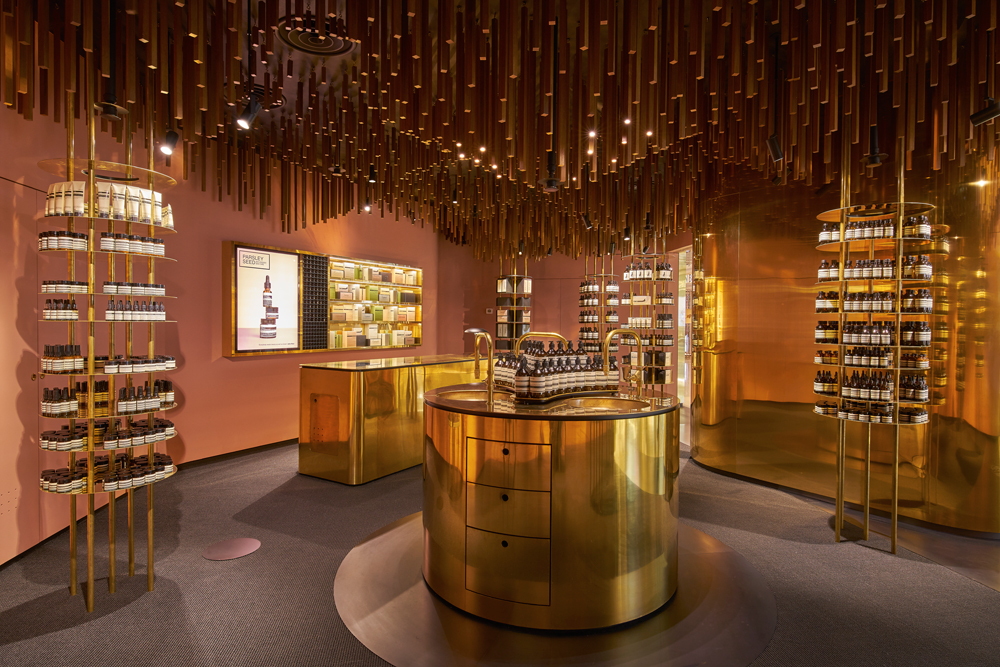 ION à Singapour est le quatrième projet réalisé par le bureau norvégien Snøhetta pour Aesop. Inaugurée en mai 2016, la luxueuse boutique en sous-sol rappelle le passé de plantation de muscade de Orchard Road. Si le laiton ou la teinte rosée des murs effleure l’histoire des lieux, le plafond se fait plus narratif et illustre les racines d’un verger au moyen d’une composition en bois.